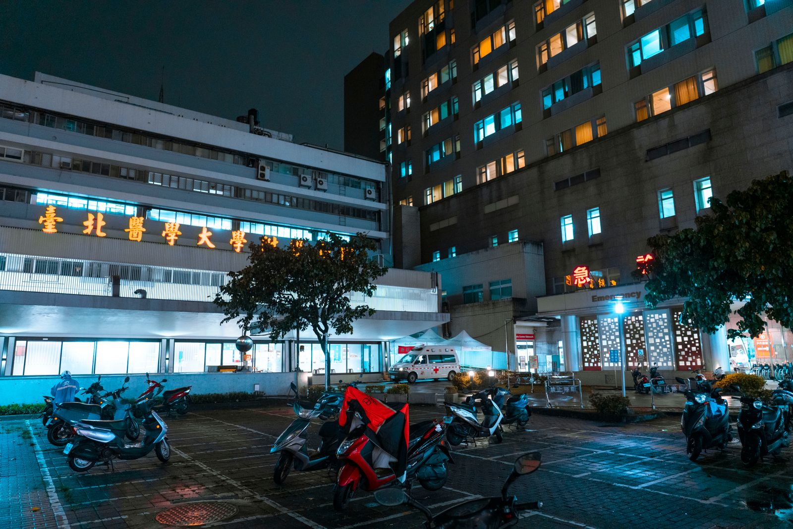 Night view of a hospital with scooters parked outside, illuminated by bright city lights.